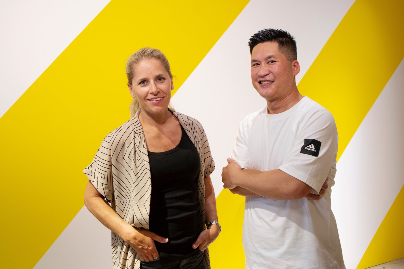 Two people stand against a yellow and white striped wall, smiling at the camera.