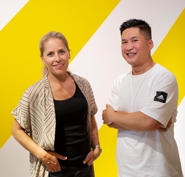 Two people stand against a yellow and white striped wall, smiling at the camera.
