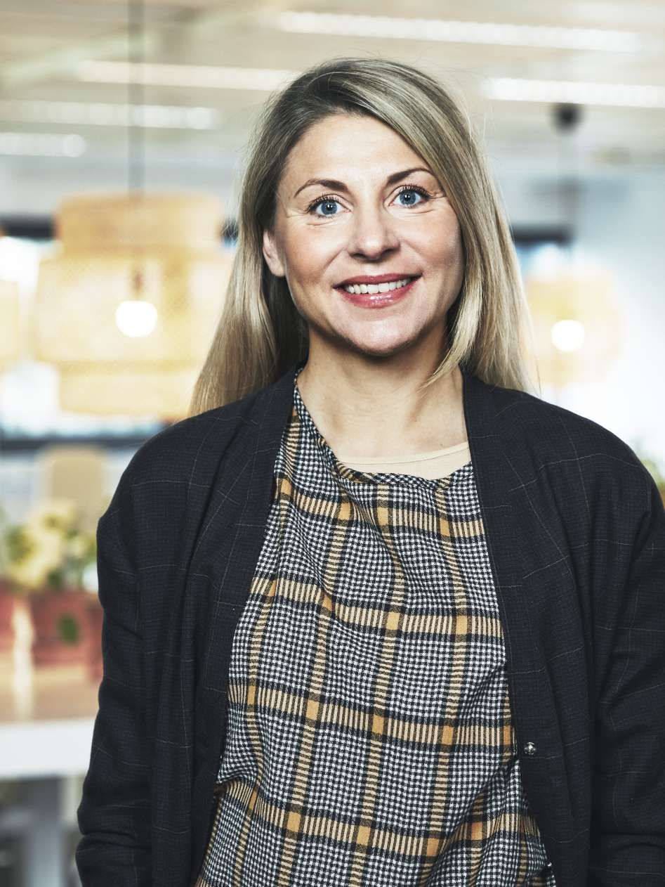 A woman smiles. She has long hair and is wearing a cardigan. There is a lamp hanging in the background.