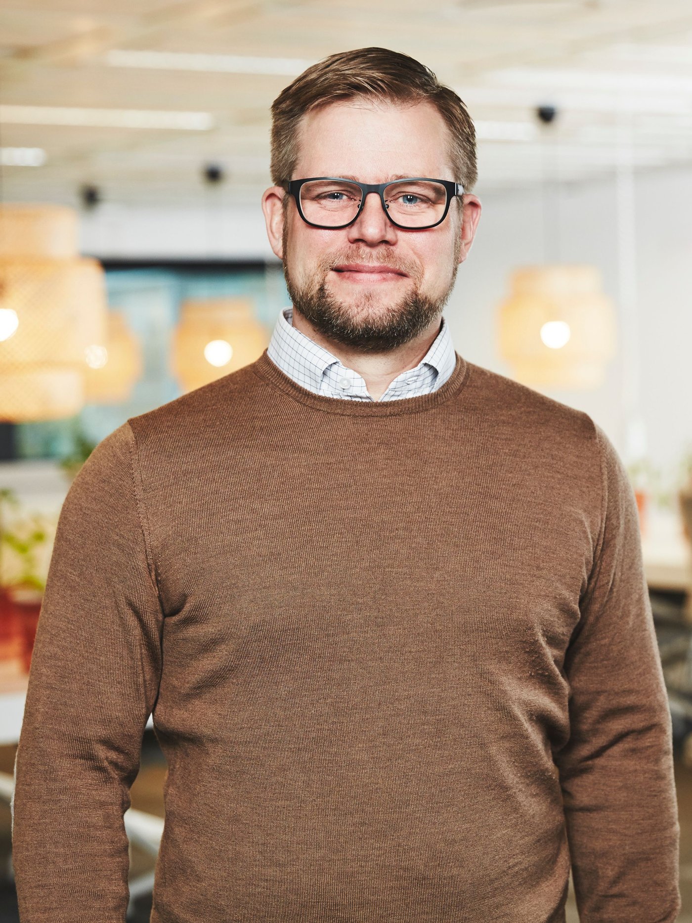 A man in a shirt and brown jumper looks and smiles at the camera. He has glasses and a beard.