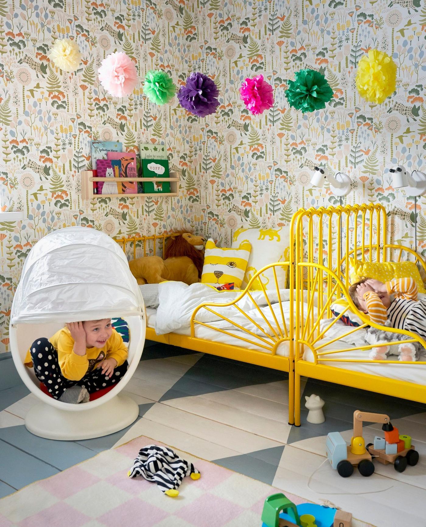 Colourful children's room with two beds place end-to-end against a wall, a a child peeking out from a white, egg-shaped swivel chair.
