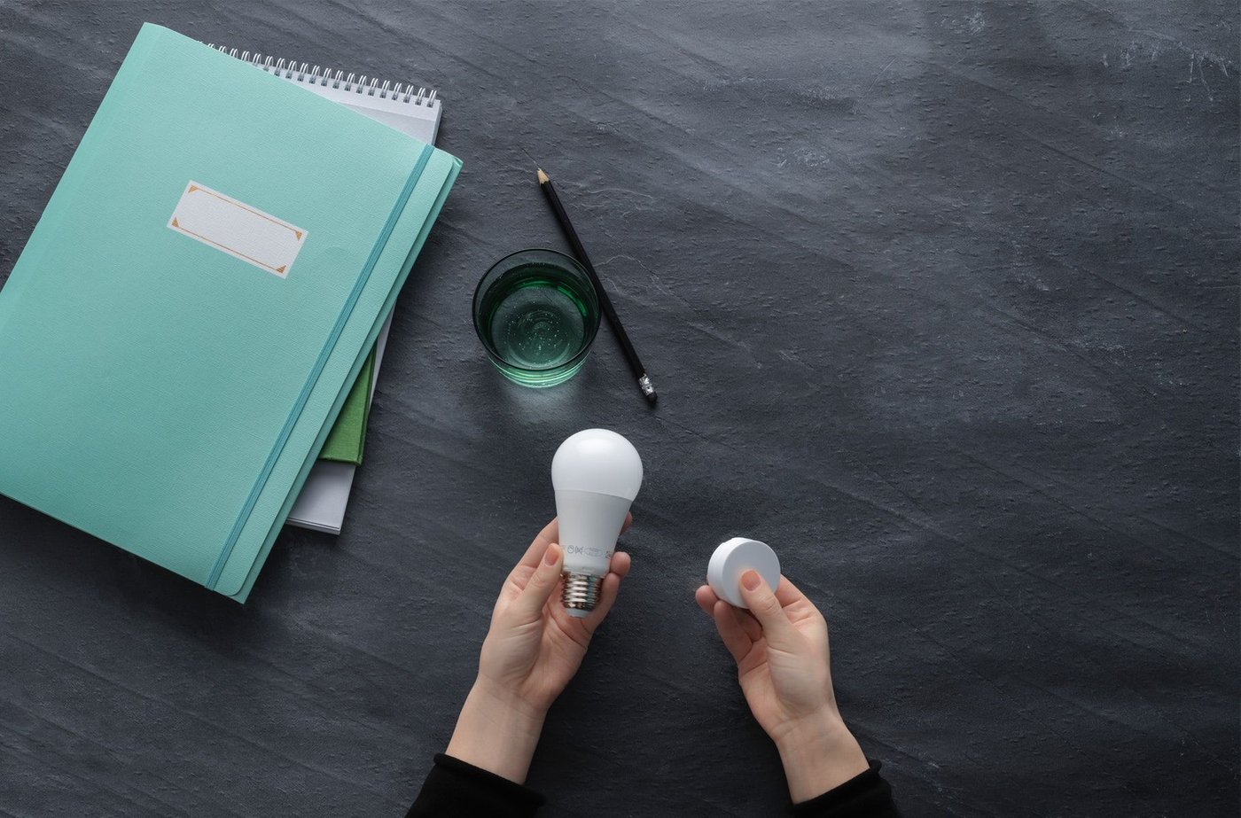 Dark grey tabletop with green notebook, a pencil and a glass, with a person's hands holding a smart lightbulb and round, white device. 