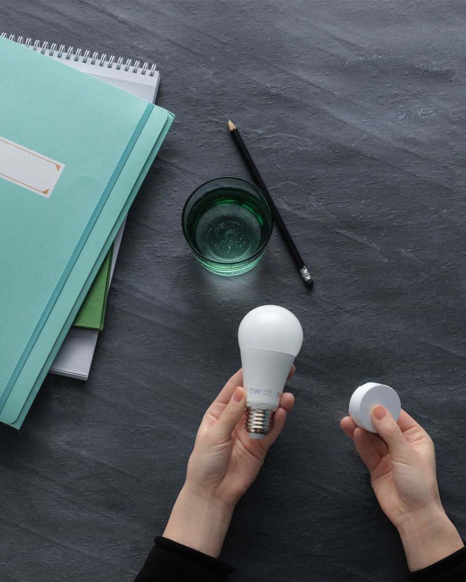Dark grey tabletop with green notebook, a pencil and a glass, with a person's hands holding a smart lightbulb and round, white device. 