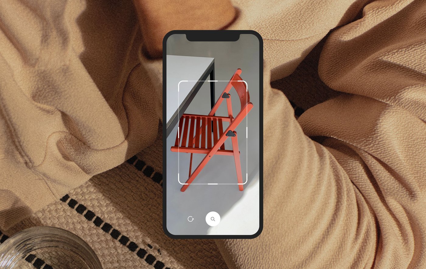 Extreme close up of a person in a beige hoodie, overlaid with a smartphone screen showing a rendering of a red folding chair being placed at a table.
