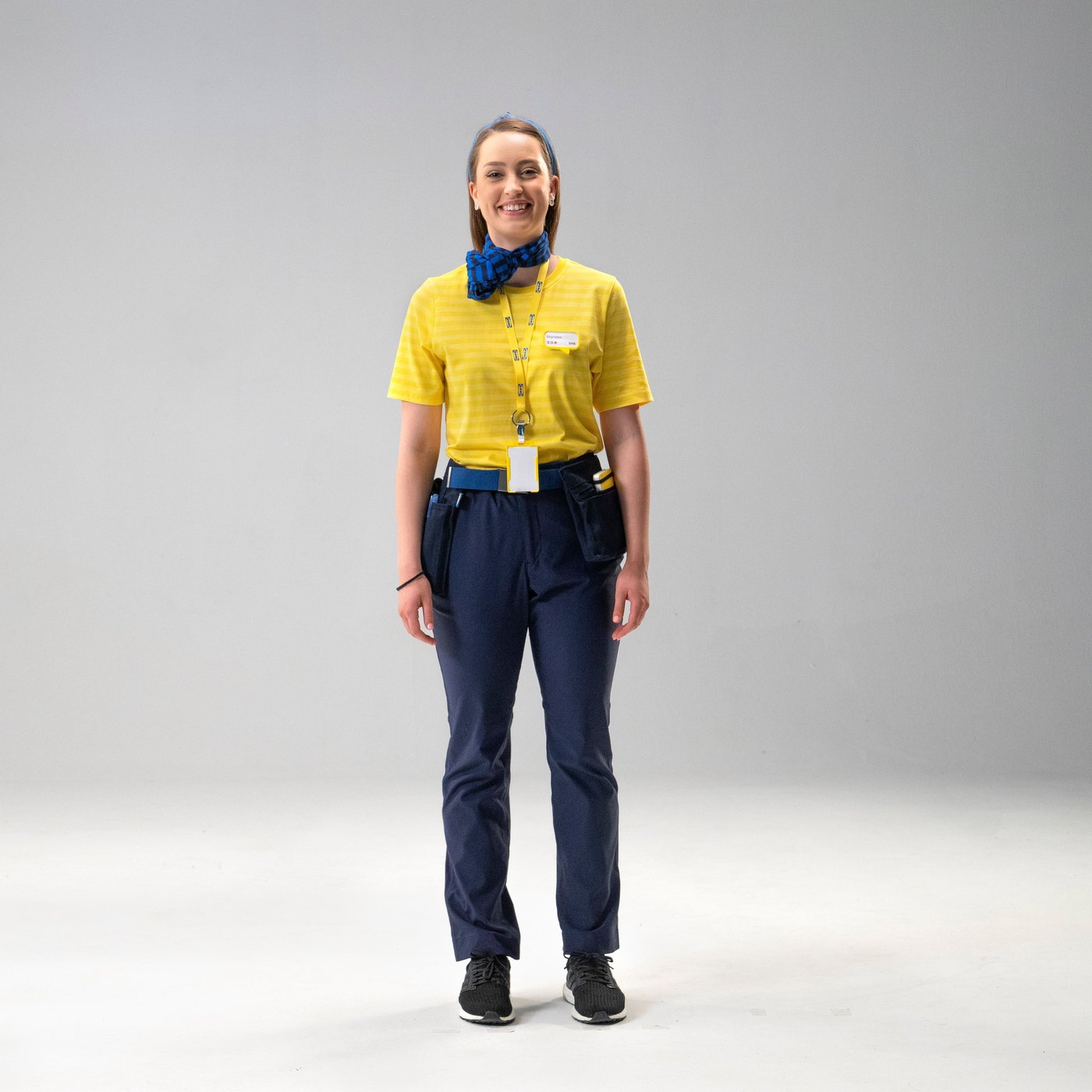 A young woman smiles at the camera. She wears blue trousers and neckscarf, a yellow  t-shirt and a lanyard.