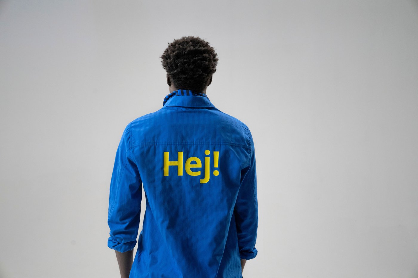 A person stands with their back to the camera. They are wearing a blue shirt with "Hej!" written on the back.