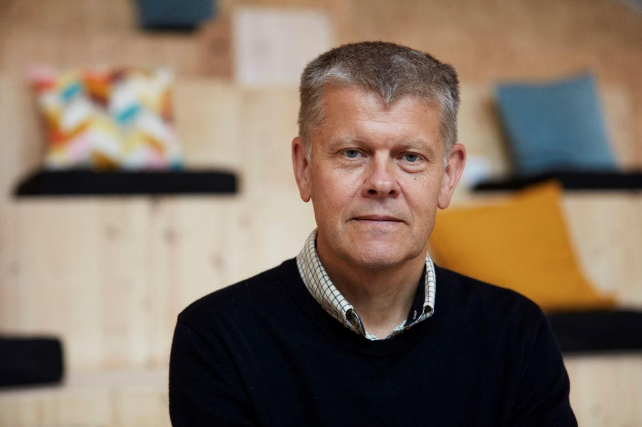 A man in a shirt and sweater stands in front of wooden steps with cushions and seating.