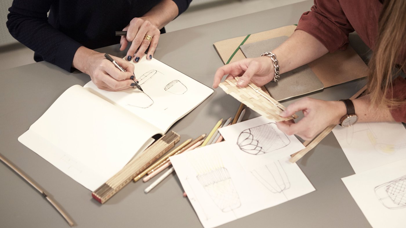 Two pairs of hands are working on a table. One person is writing, the other is looking at a piece of wood. There are papers and pencils on the table.