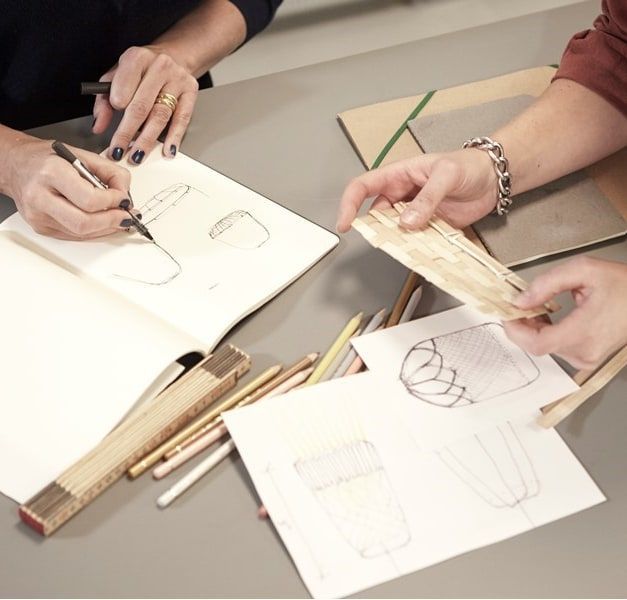Two pairs of hands are working on a table. One person is writing, the other is looking at a piece of wood. There are papers and pencils on the table.