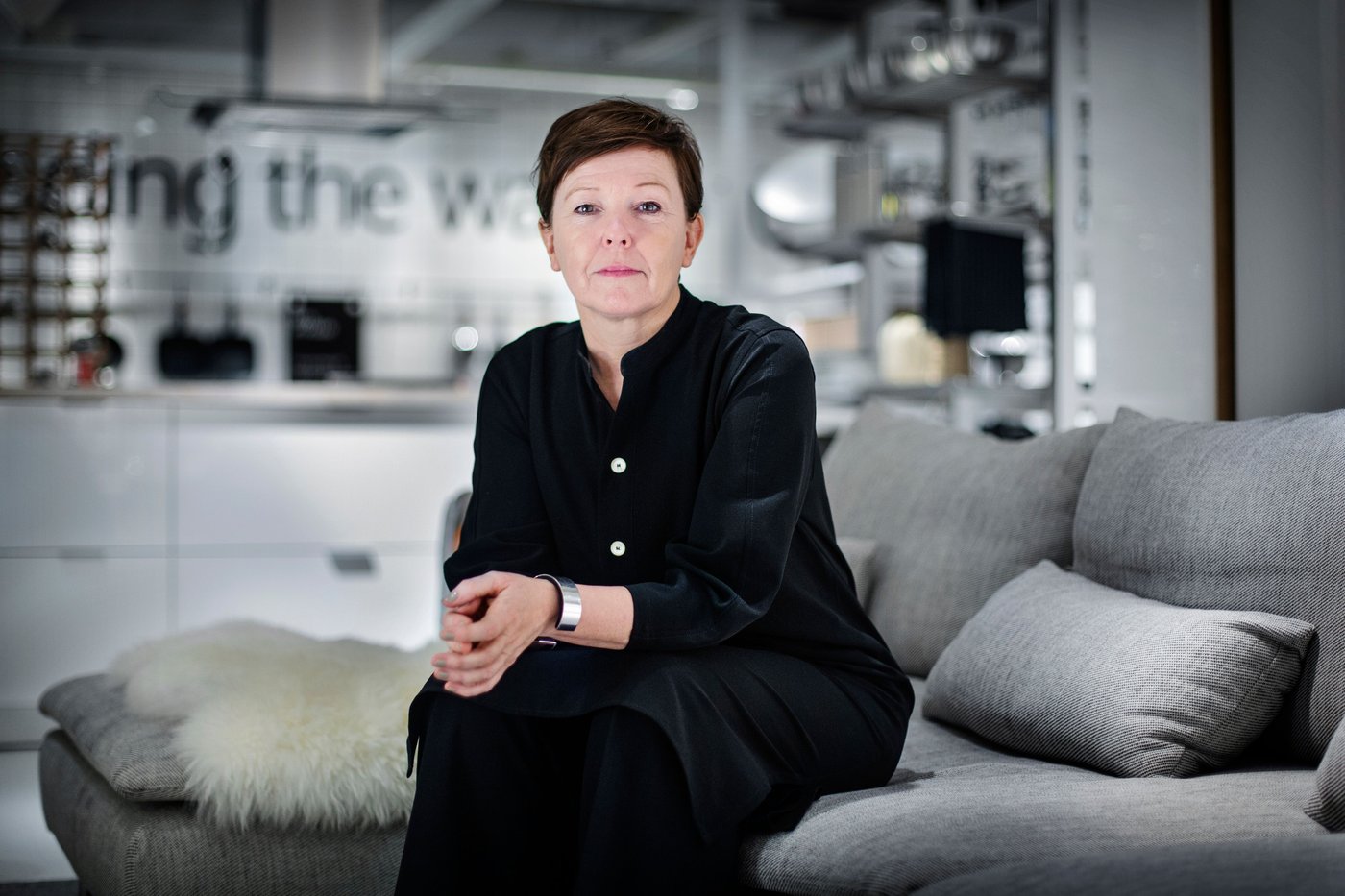 A woman wearing black sits on a sofa. There is a kitchen behind her.