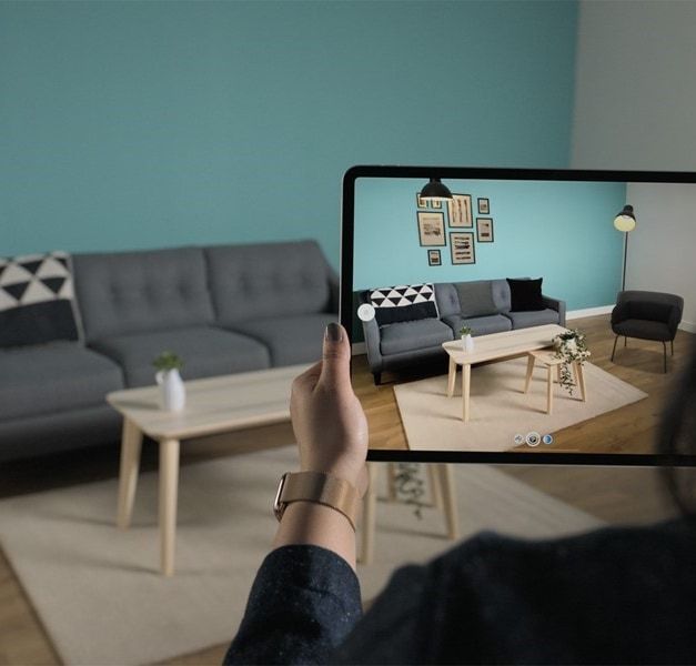 A person points an iPad screen at a grey sofa and wood table in a room. The wall is blue/green.