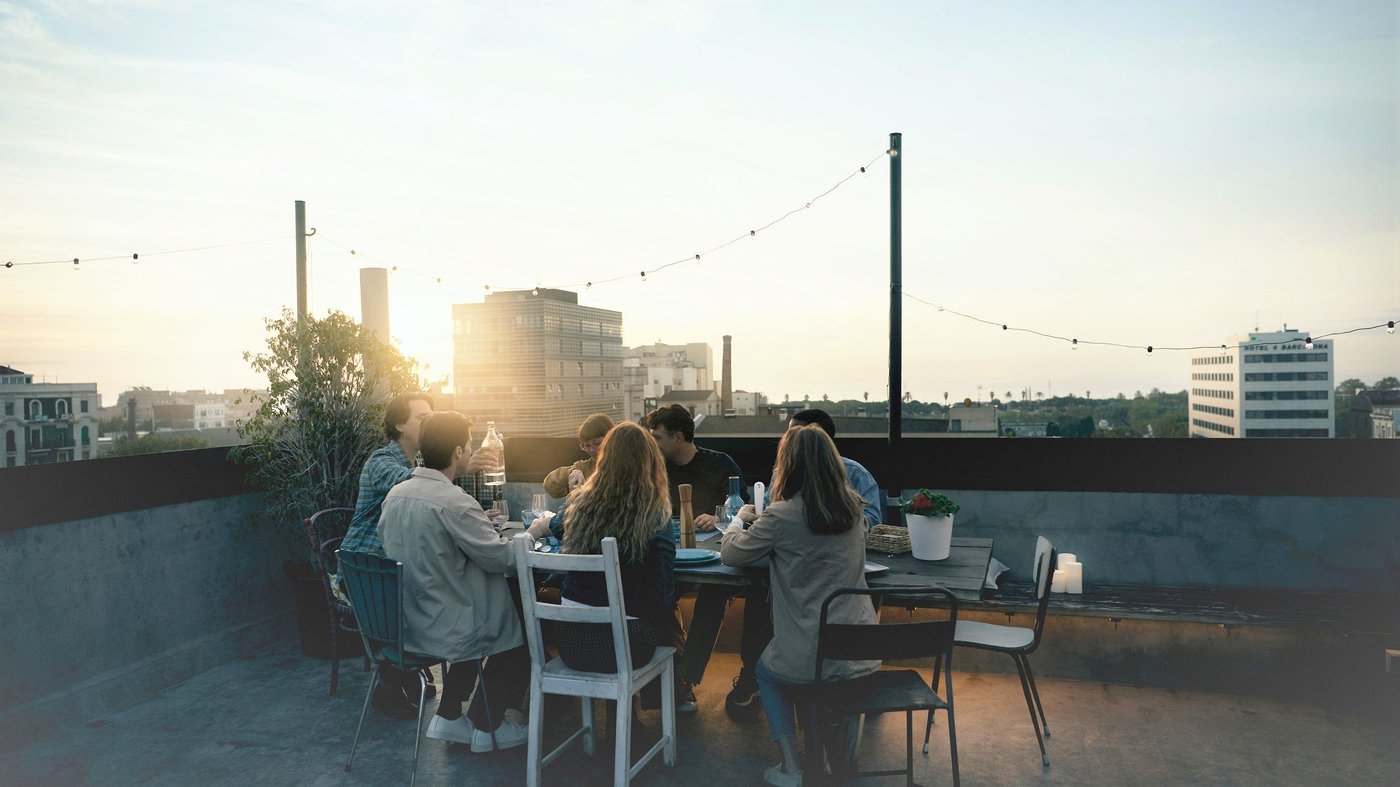 A group of people sit around a table on a roof terrace. The sun is shining in the background.