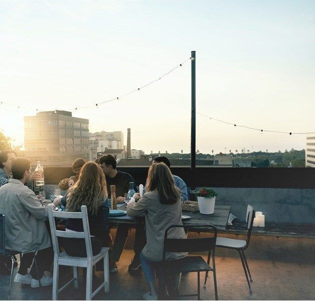 A group of people sit around a table on a roof terrace. The sun is shining in the background.