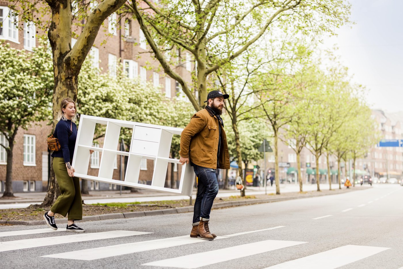 A woman and a man carry a white shelving unit across a zebra crossing on a tree-lined road.