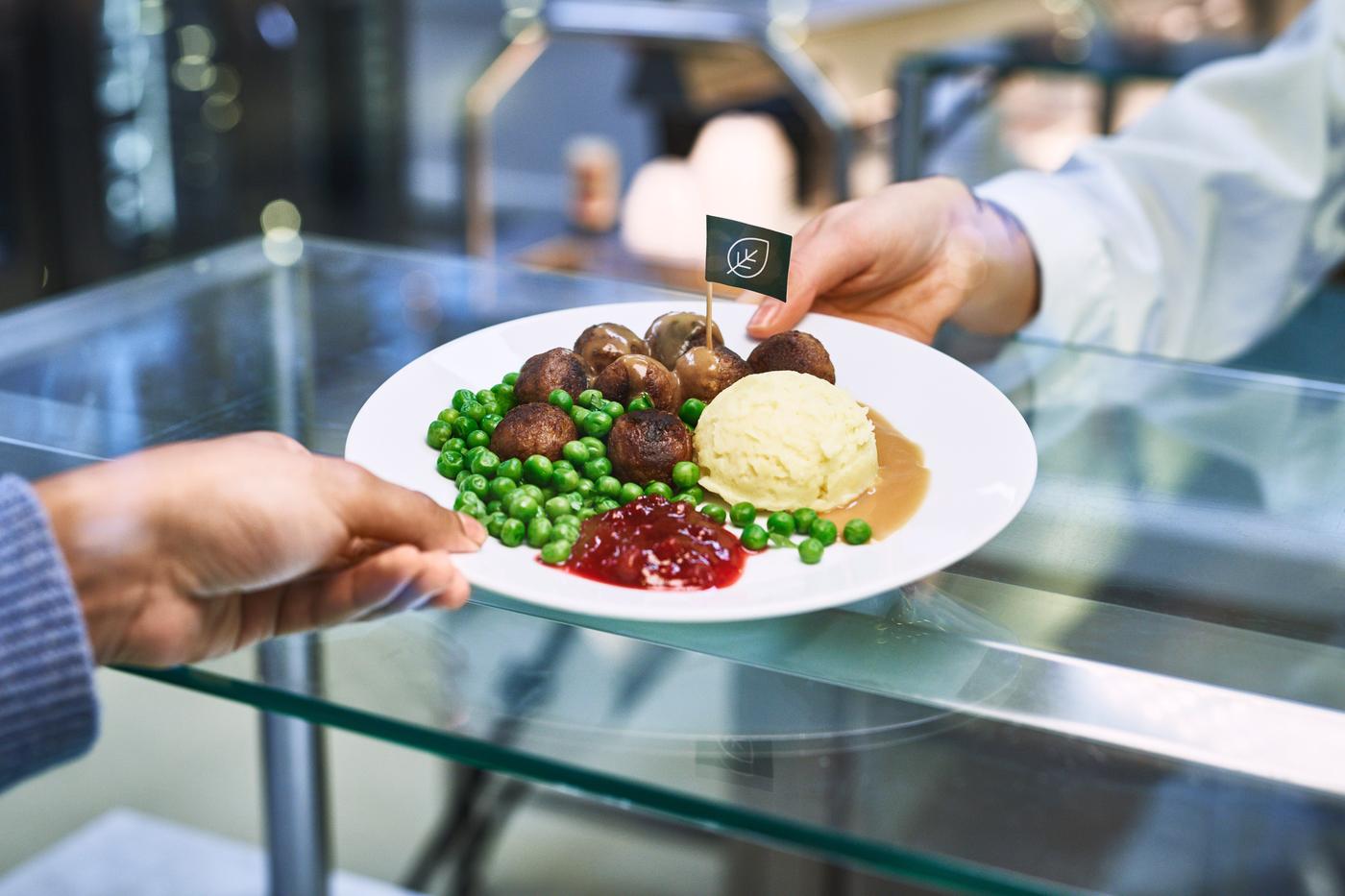 A person hands over a plate with meatballs, mashed potato, peas and lingonberry sauce to another person, over the counter.
