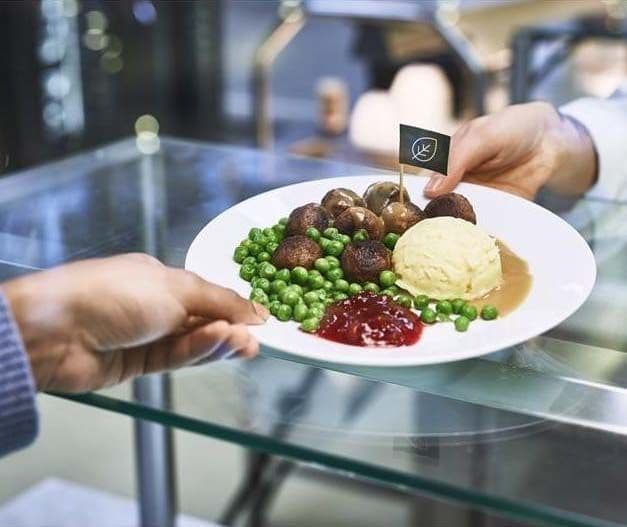 A person hands over a plate with meatballs, mashed potato, peas and lingonberry sauce to another person, over the counter.