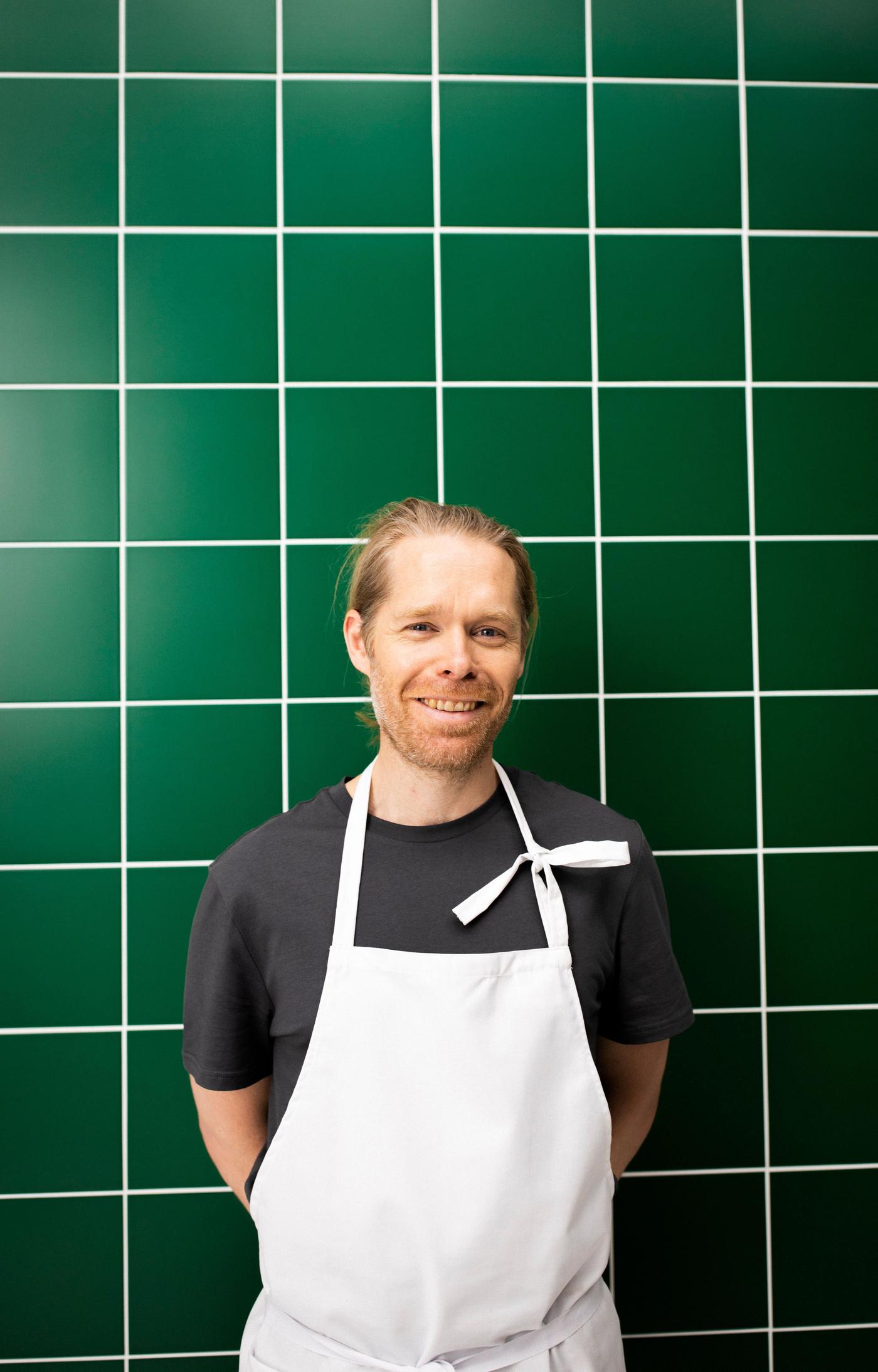 A man stands in front of a green tiled wall. He is smiling. He wears a white apron.