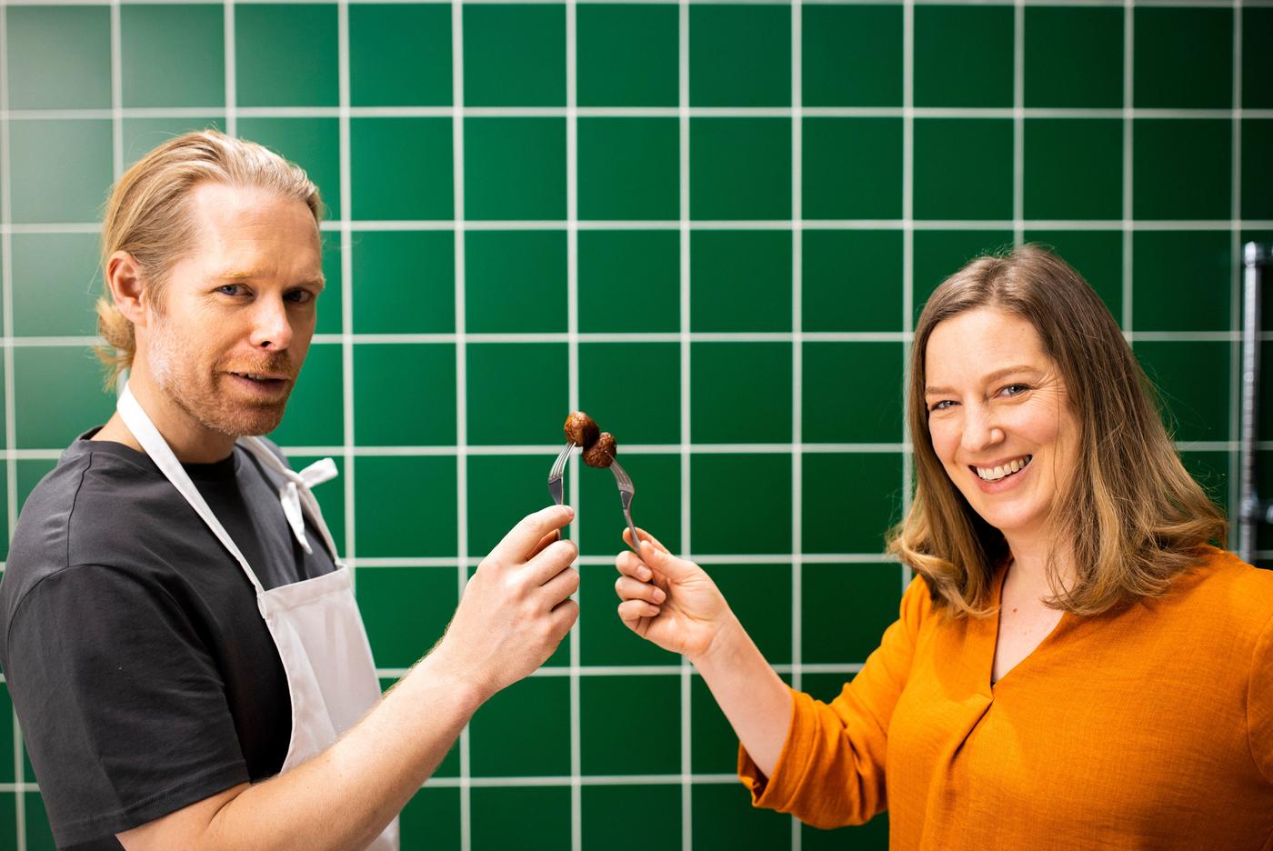 A man and woman stand in front of a green tiled backdrop. They are smiling and each is holding a meatball on a fork.