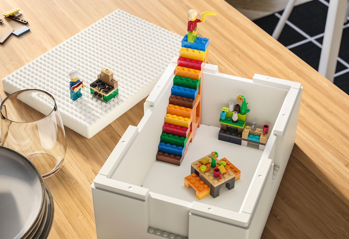 A white storage box containing lego blocks and characters stands on a wood table. There is a plate and a glass next to it.