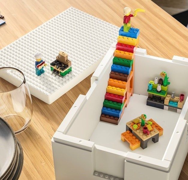 A white storage box containing lego blocks and characters stands on a wood table. There is a plate and a glass next to it.