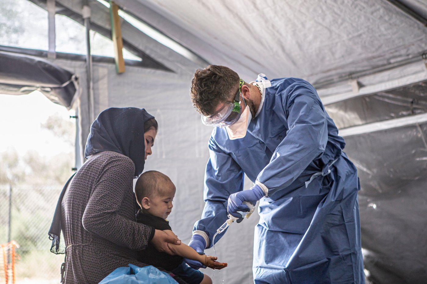 A medical professional in blue protective wear treats a child while it sits on a woman's lap.