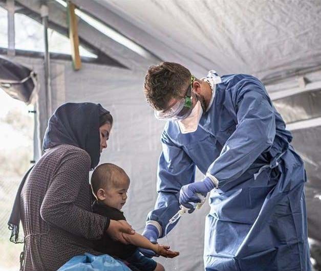 A medical professional in blue protective wear treats a child while it sits on a woman's lap.