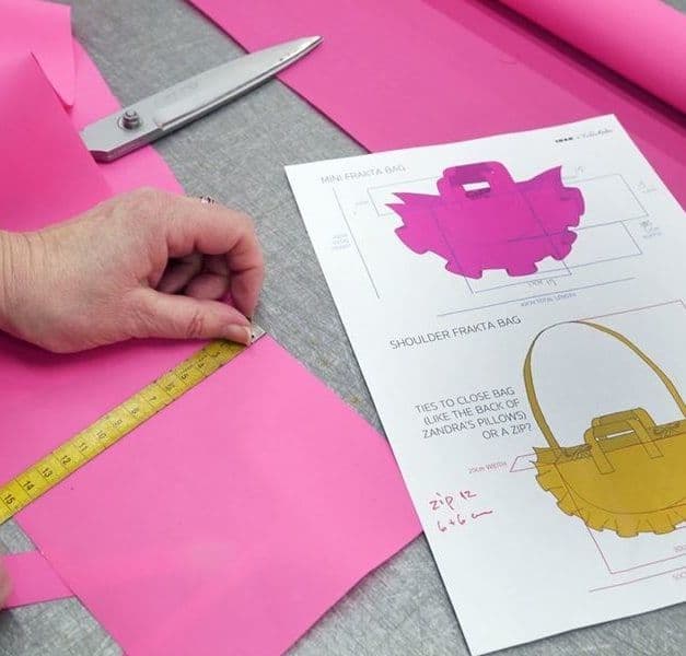 A pair of hands measure a piece of pink fabric, following a bag pattern. There are a pair of scissors too.