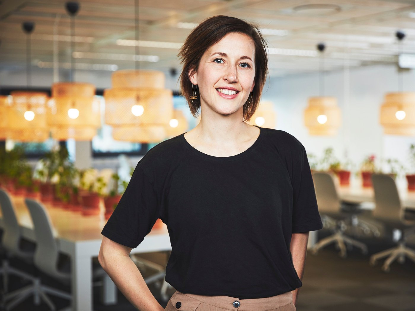 A woman in a black top stands and smiles at the camera. She is in a large office with desks, chairs and pendant lamps behind.