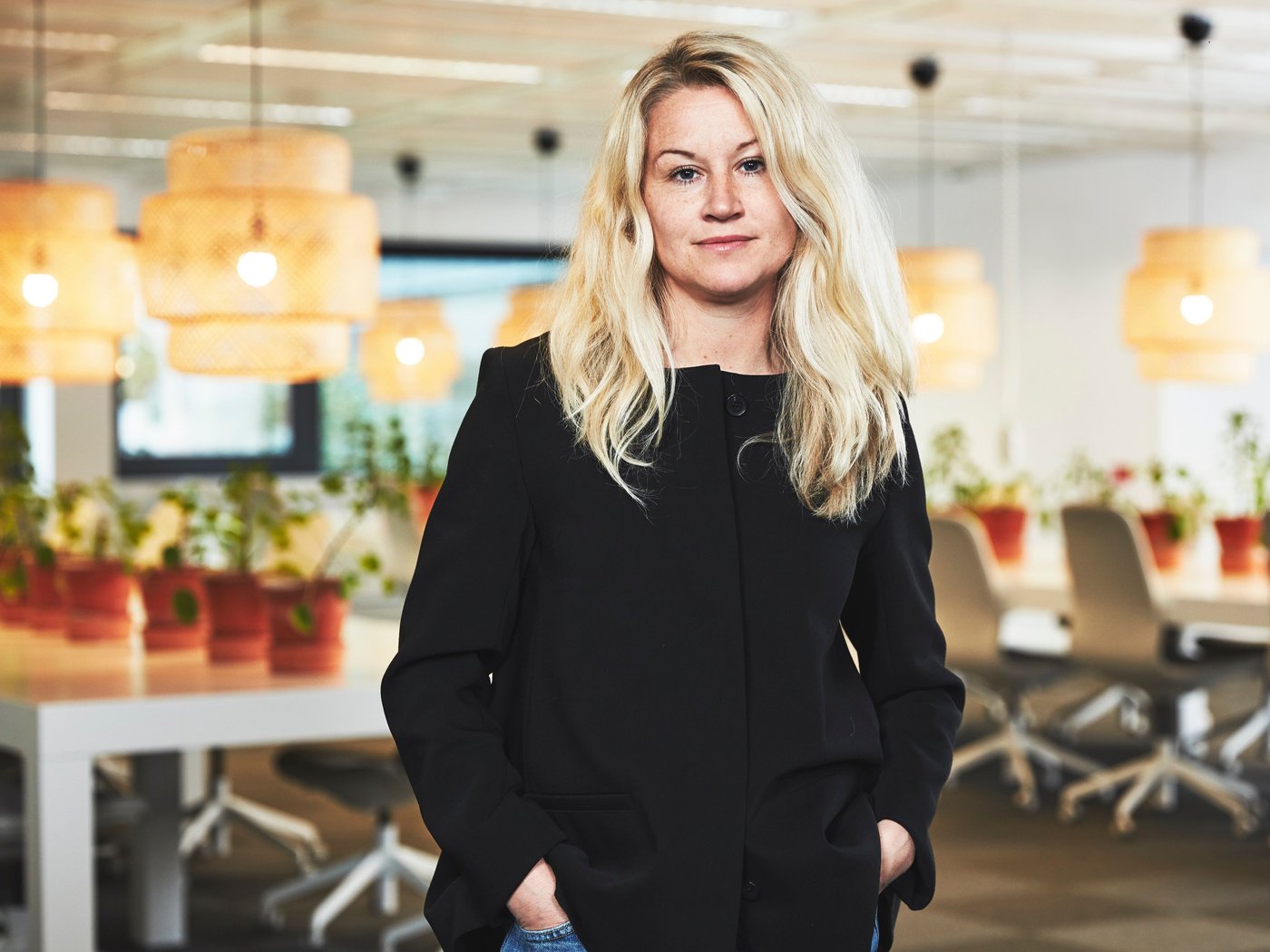 A woman wearing black stands in a large office. There are desks and lamps behind her.
