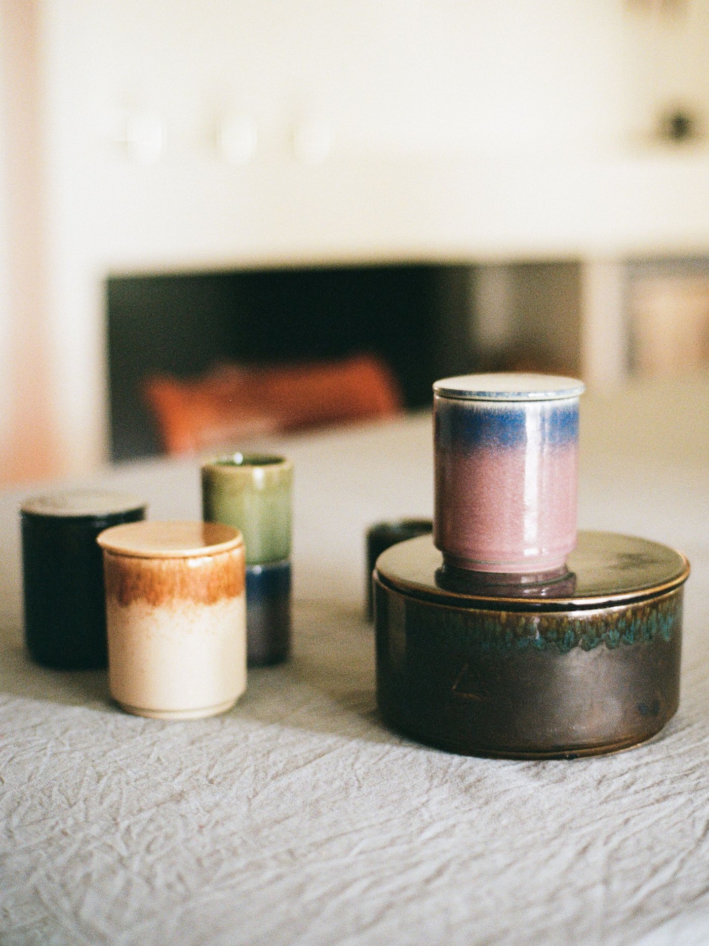 Three ceramic vessels holding candles stand on a wood table. Another sits on top of a ceramic container.