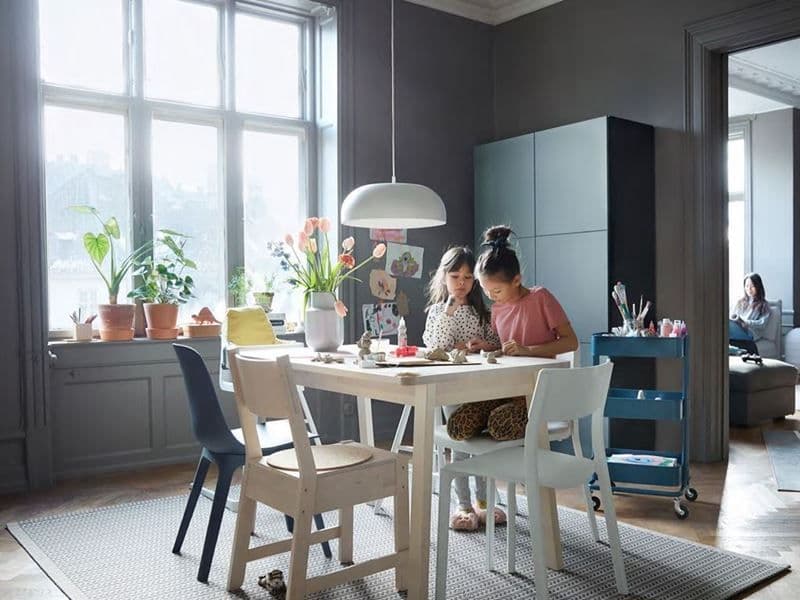 A child sits at a table, while another stands and looks over her. They are drawing in the dining area. A woman sits in the room next door, reading.