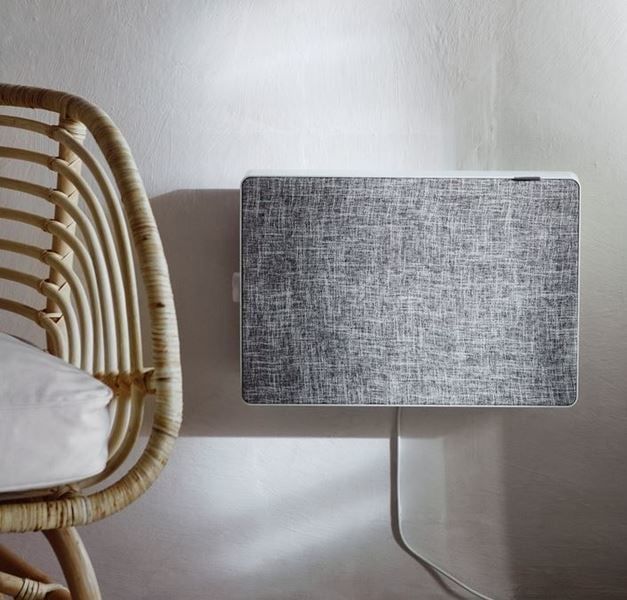 A grey air purifier is fixed to a pale wall. To the left is a chair and on the right is a white curtain.