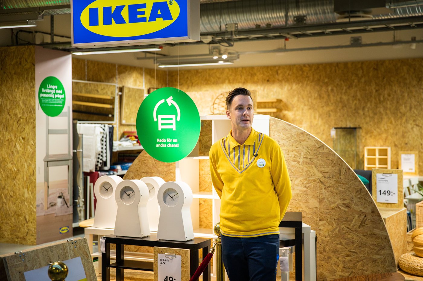 A man stands in front of an IKEA secondhand store. There are four white clocks behind him and a large green sign.