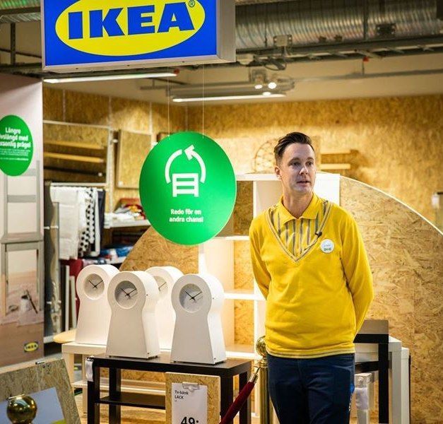 A man stands in front of an IKEA secondhand store. There are four white clocks behind him and a large green sign.