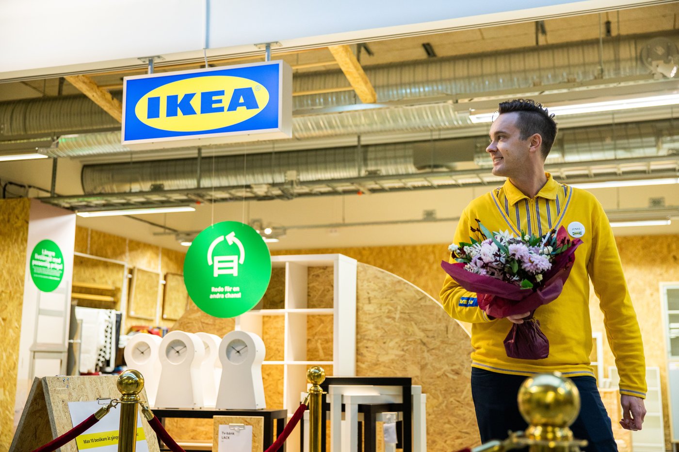 A man is walking in front of the IKEA secondhand store. He has a bunch of flowers in his hand. He is smiling.