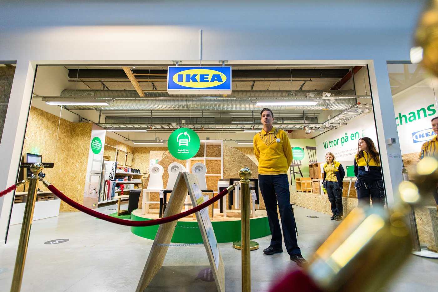 Four IKEA co-workers stand in the IKEA secondhand store. There is an IKEA sign above and a velvet rope in the foreground. There are lots of products, but no customers yet.