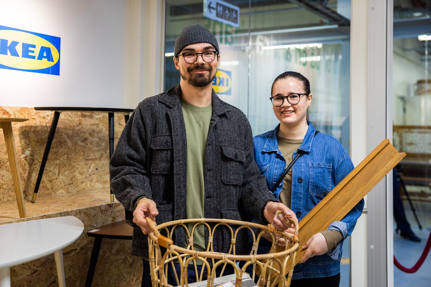 A man and woman smile at the camera. The man is carrying a bamboo basket. The woman is holding a piece of wood. 