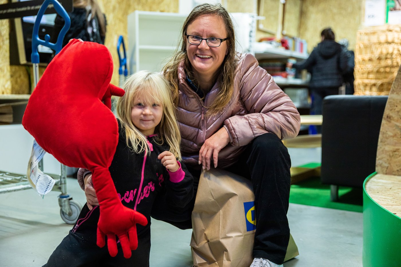 A woman and child kneel in front of the camera outside the IKEA secondhand store. The woman is smiling. The child holds a large heart cushion.