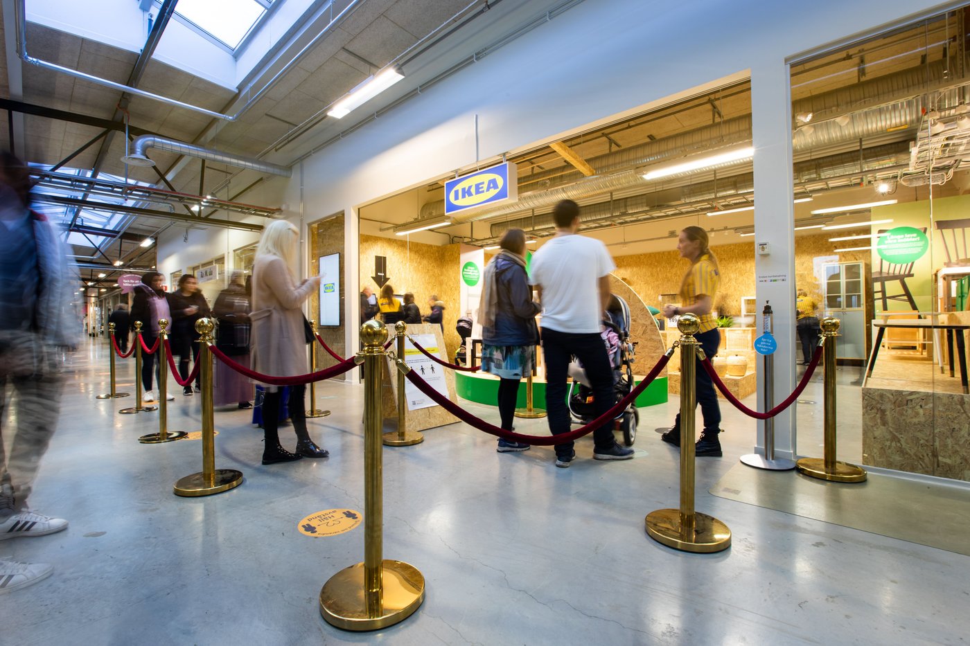 Customers enter the IKEA secondhand store. They walk through the red ropes. They have their backs to the camera. There are more people waiting to enter.