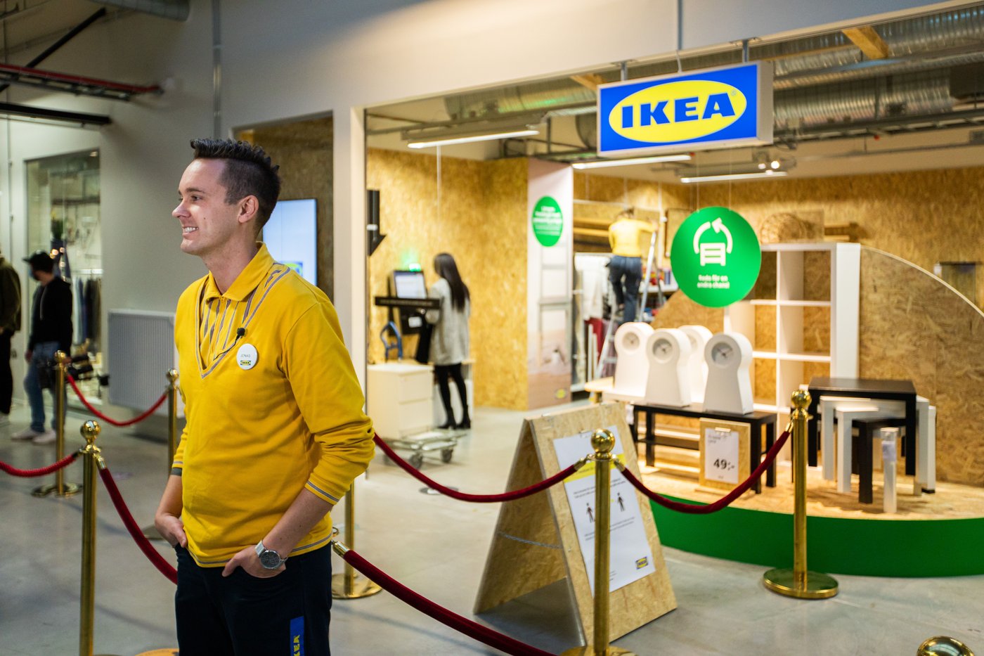 An IKEA co-worker stands at the front of the IKEA secondhand store. He stands in front of the red velvet ropes. He looks at the cameras of the photographers.