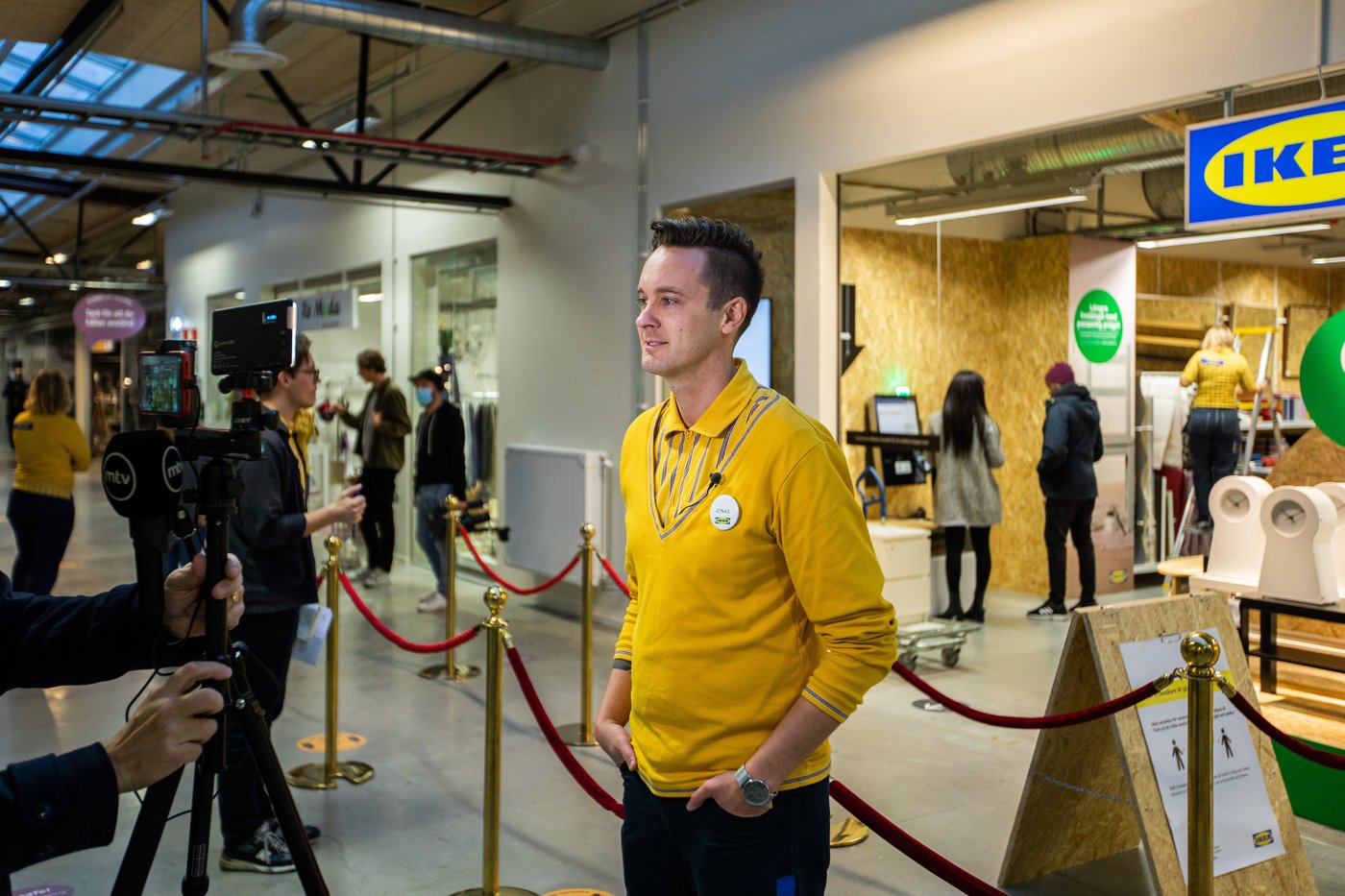 An IKEA co-worker stands at the front of the IKEA secondhand store. He stands in front of the red velvet ropes. He looks at the cameras of the photographers.