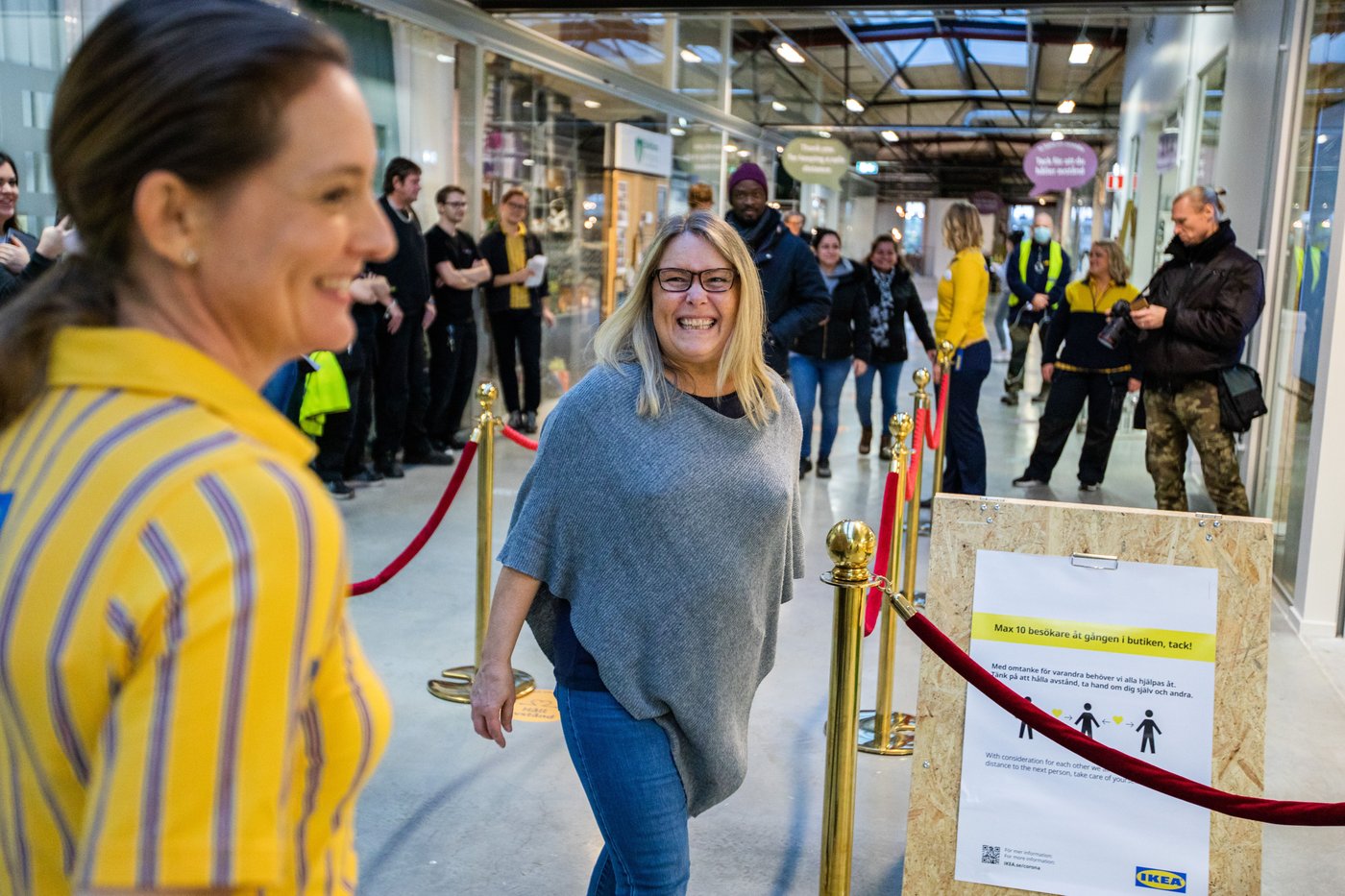 A woman laughs as she walks through the velvet ropes into the IKEA store. To the left, an IKEA co-worker smiles. There are people standing in the background.