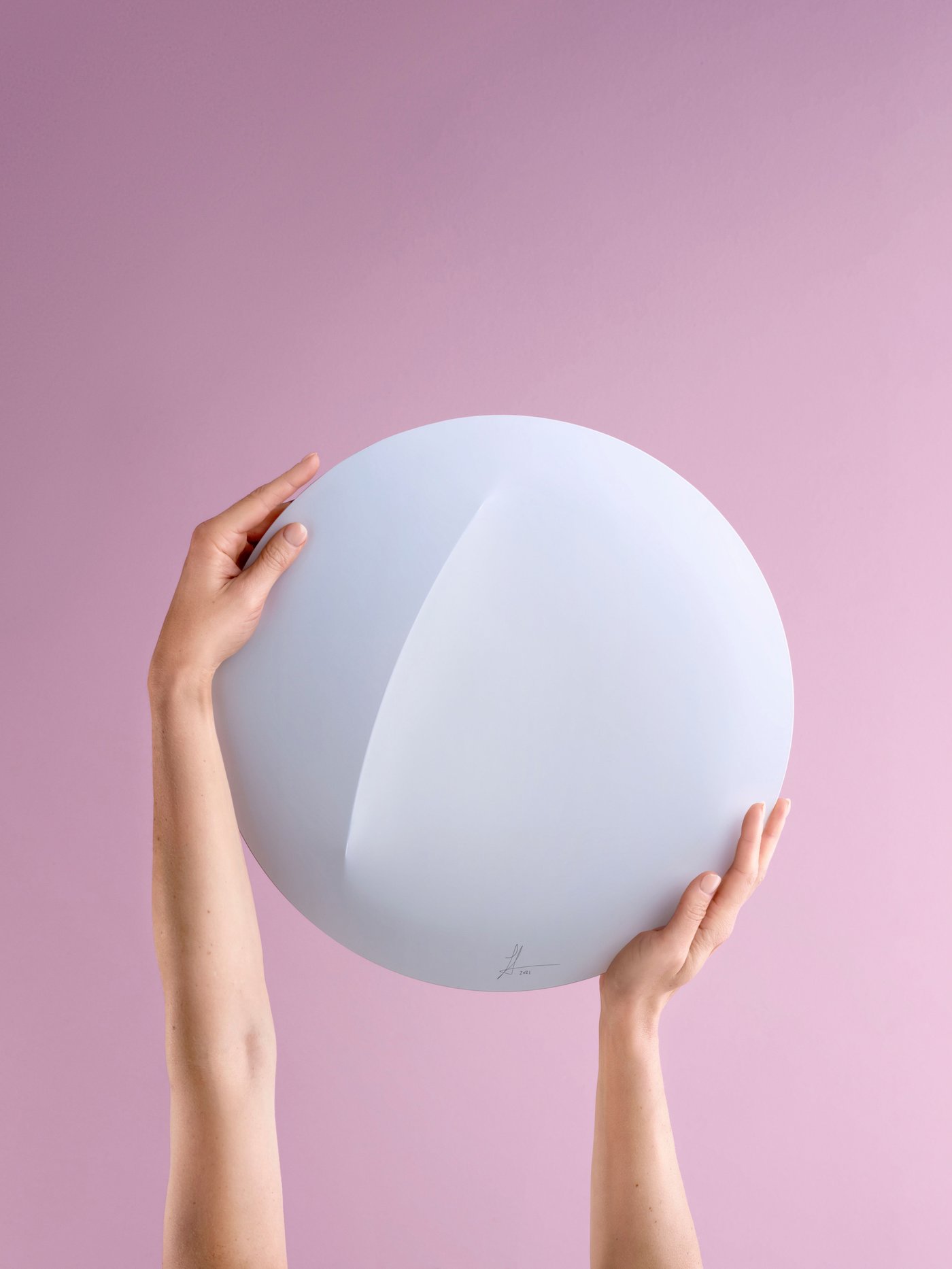 A pair of hands hold a white circular lamp against a pink background.