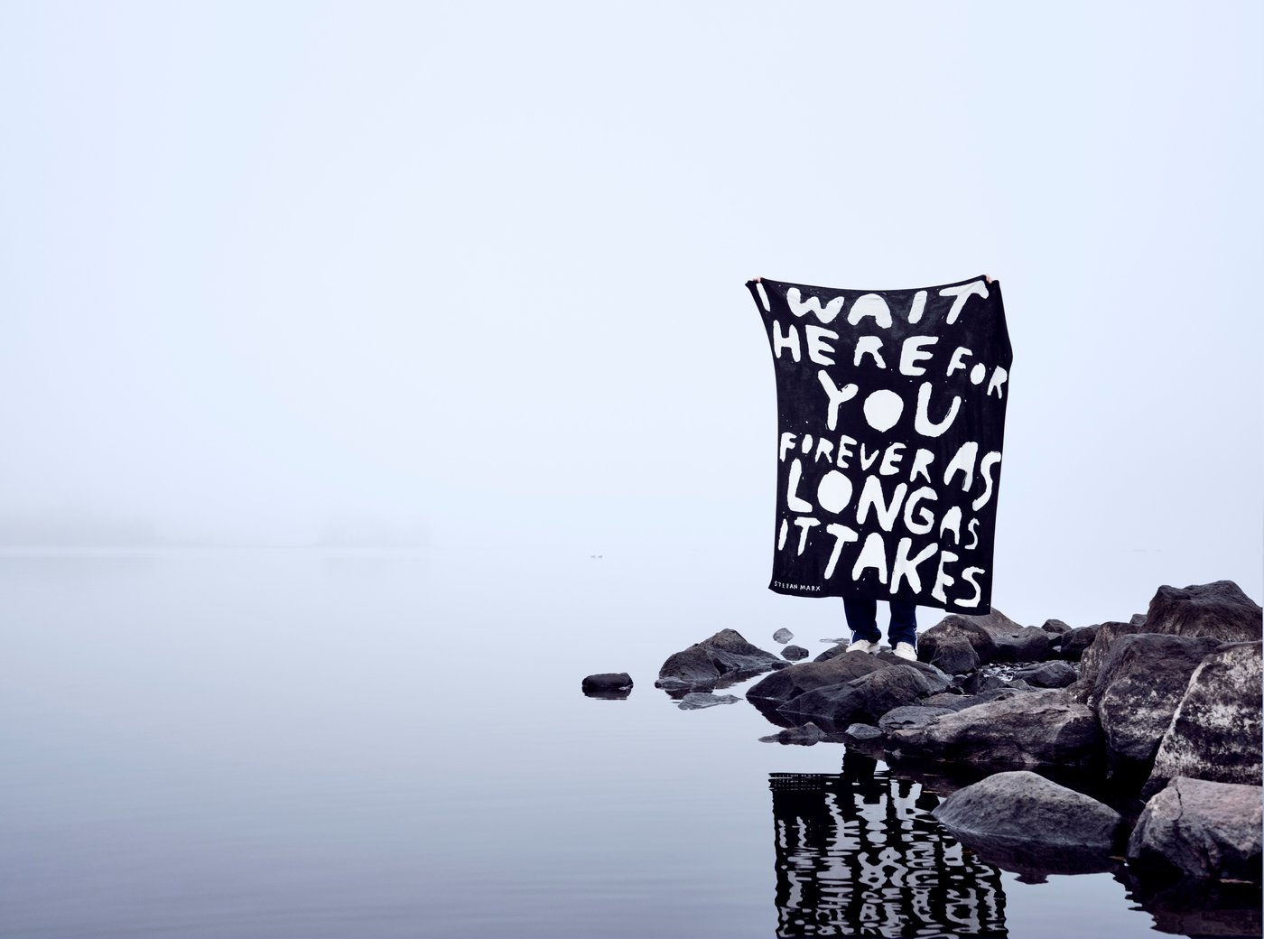 A person standing on rocks next to the water holds a large black and white throw with writing.