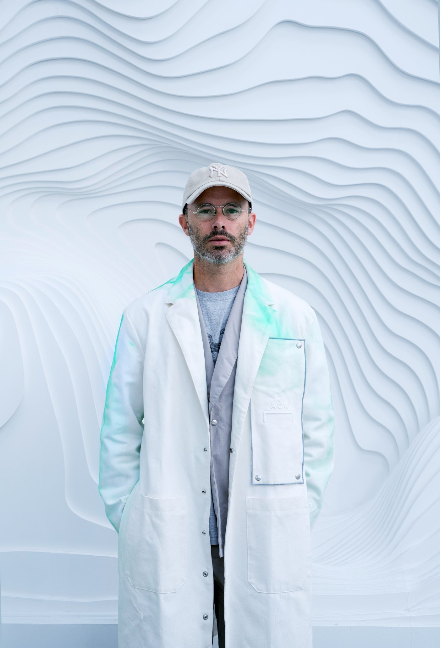 A man in a white coat and hat stands in front of a wall with an undulated design.