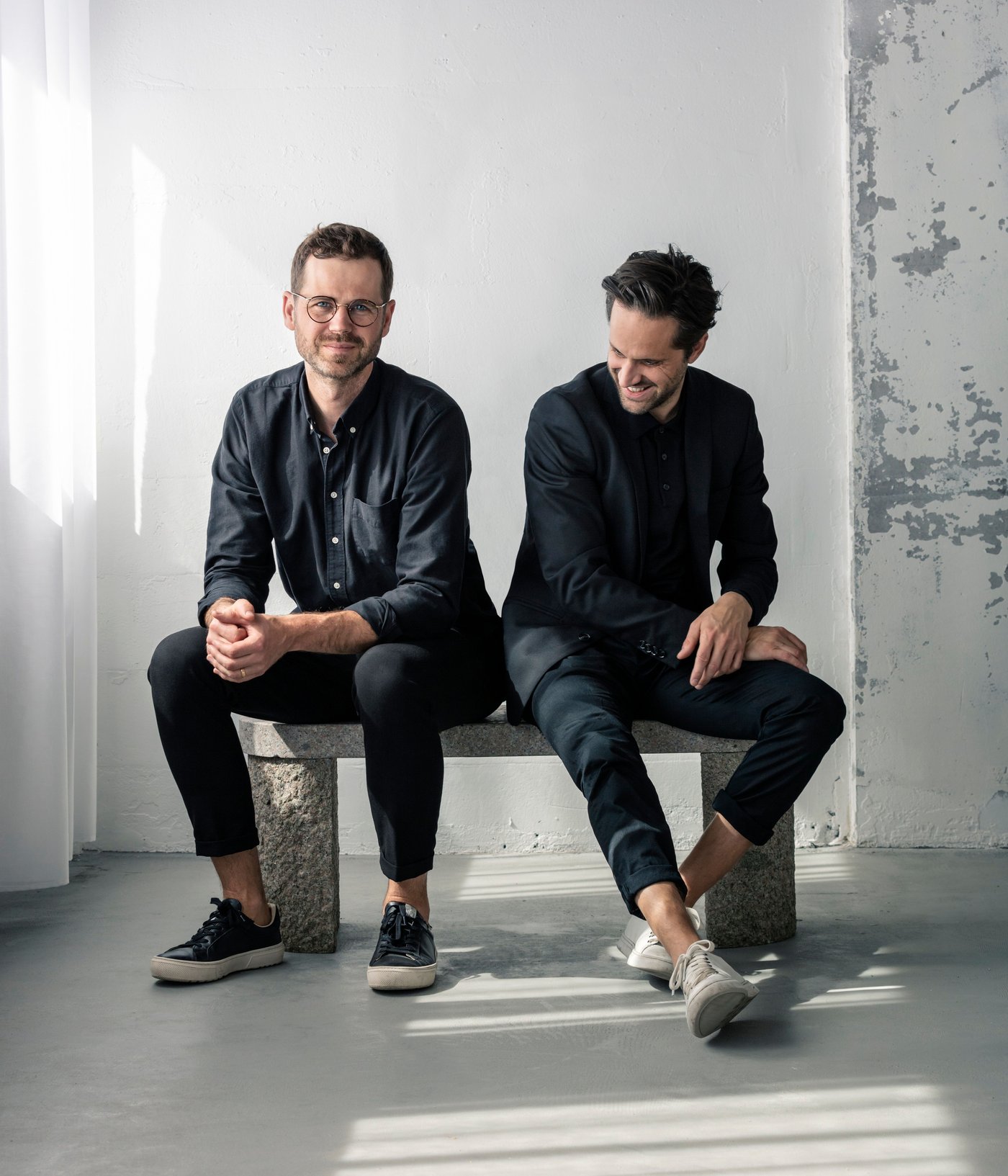 Two men sit on a bench in a white, sun filled room.