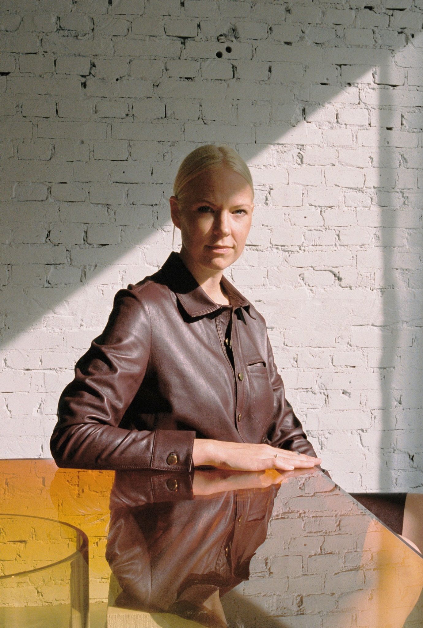 A woman wearing brown sits at a yellow glass table. The sun shines on her.