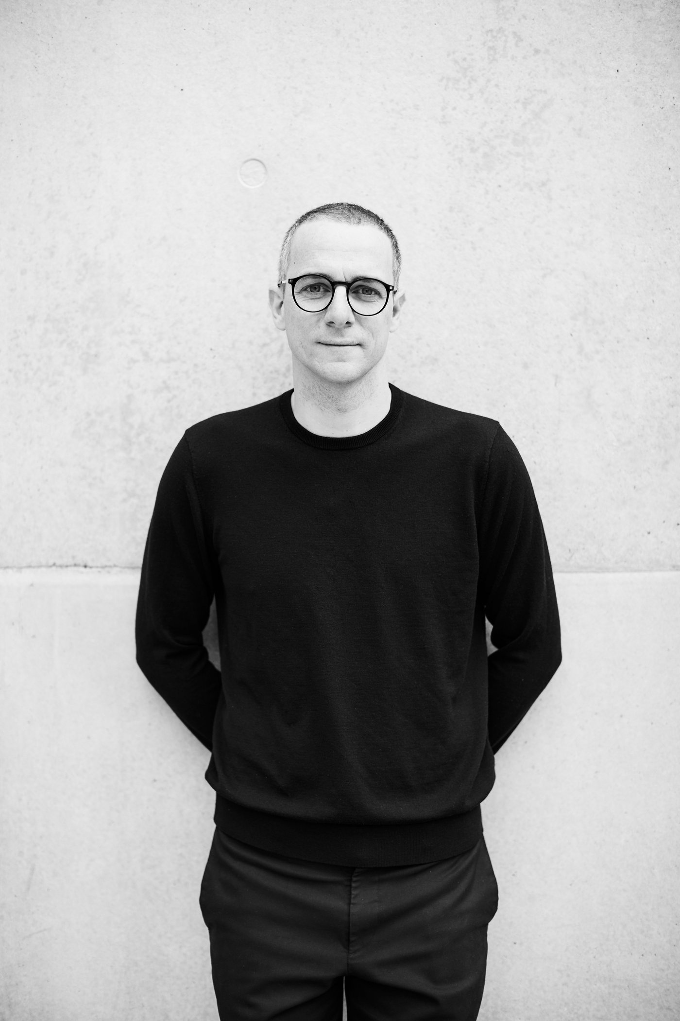 A black and white photo of a man in dark clothing standing against a wall.