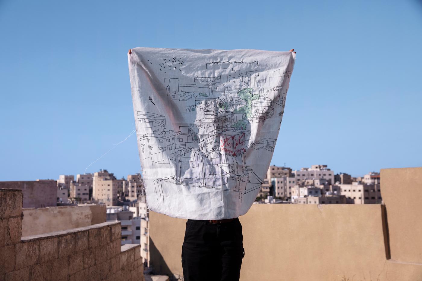 A person holds up a white fabric square with a print on it. In the background there are buildings and the sky.