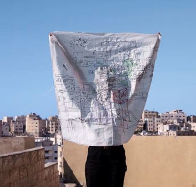 A person holds up a white fabric square with a print on it. In the background there are buildings and the sky.